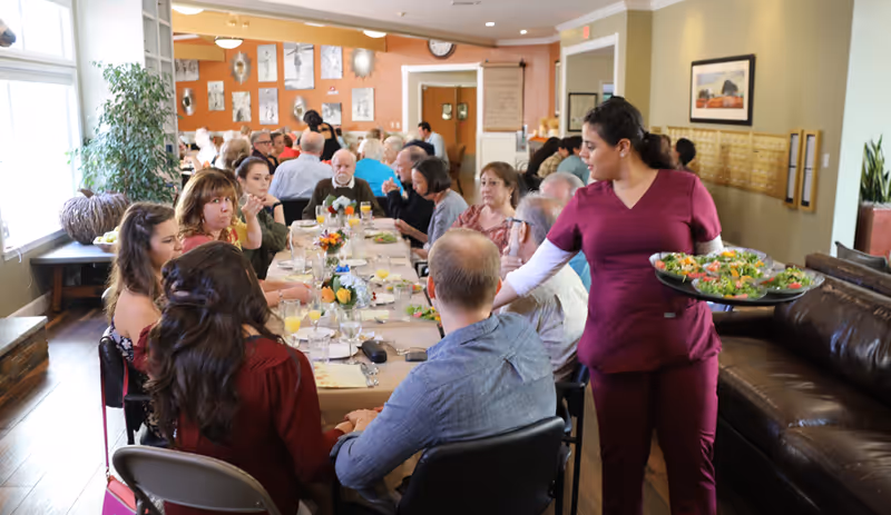 A group of elderly people and caregivers seated around a long dining table in a well-lit room, with a caregiver in maroon scrubs serving plates of salad. The table is set with glasses, plates, and floral centerpieces, and the room has large windows, plants, and warm-colored walls.