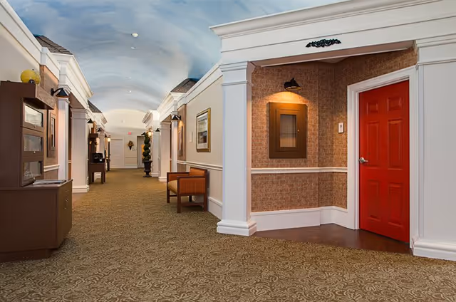 A well-lit hallway in a senior living facility with patterned carpet, white walls with decorative molding, framed artwork, and a red door on the right side. The ceiling is painted to resemble a blue sky with clouds. There are benches and decorative plants along the hallway.
