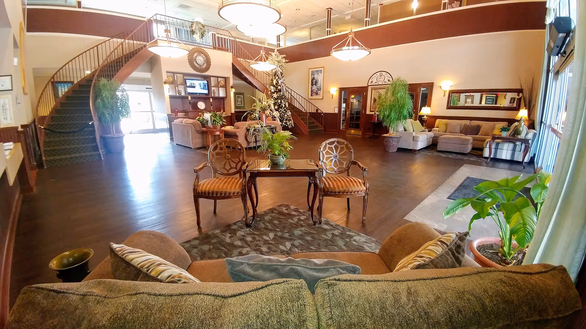 Spacious senior living community common area with multiple seating arrangements including sofas and chairs, a central table with plants, a decorated Christmas tree, large windows allowing natural light, and a staircase leading to an upper level.