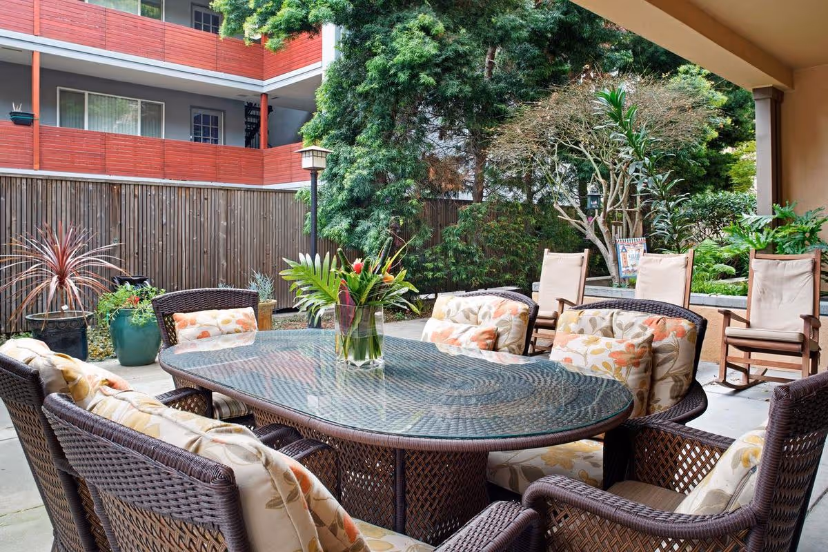 Covered outdoor patio courtyard with a glass-topped wicker dining table and cushioned chairs surrounded by plants.