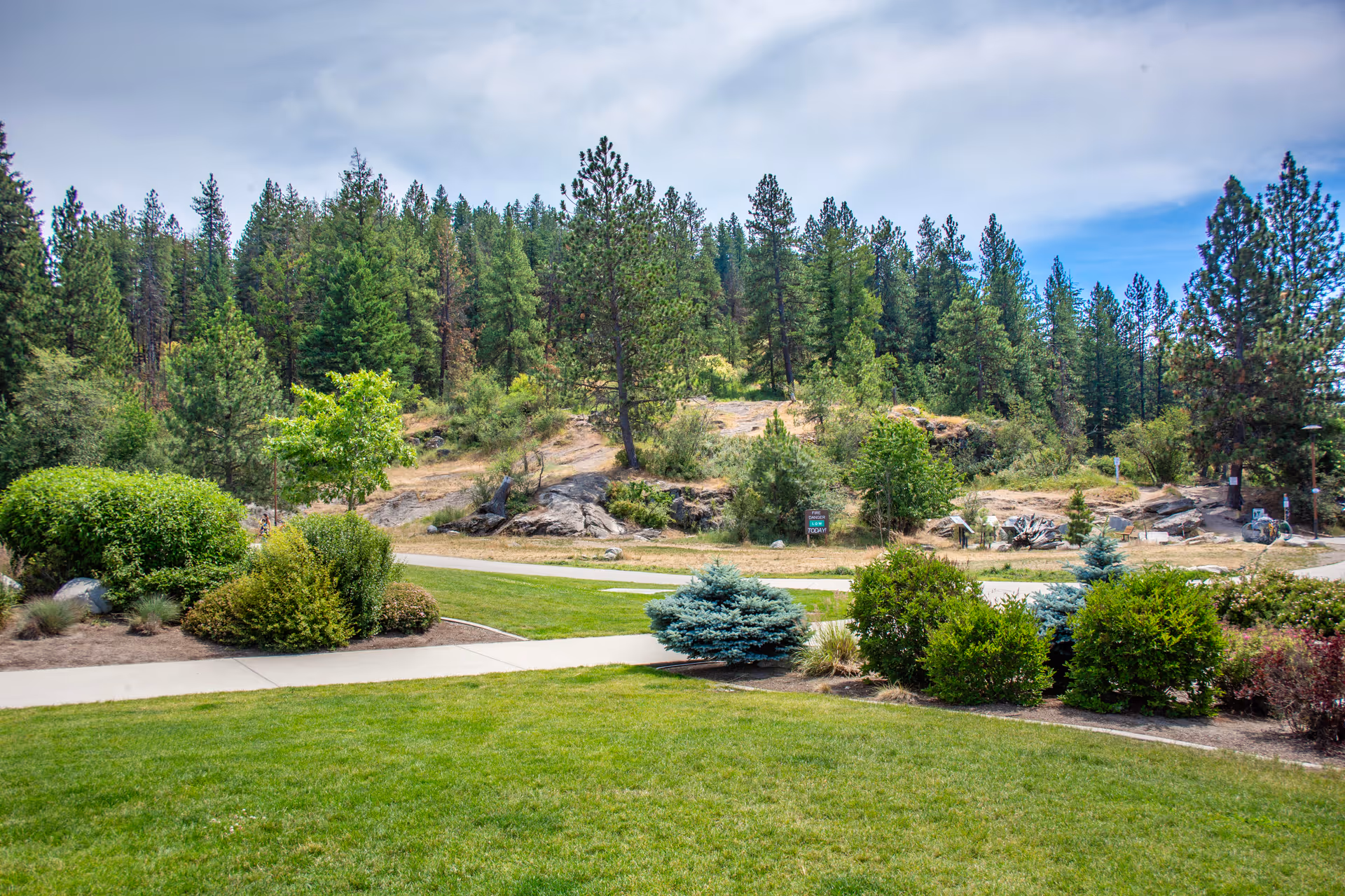 A landscaped outdoor area with a well-maintained green lawn, various bushes, and small trees. In the background, there is a forested hillside with tall pine trees under a partly cloudy sky.
