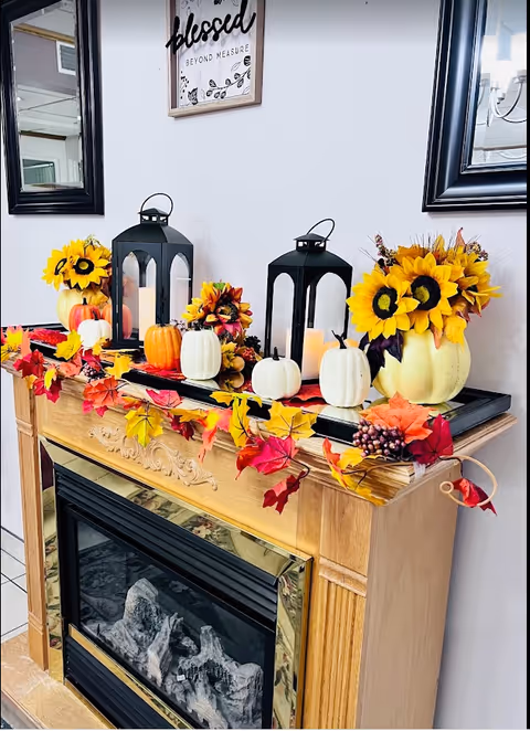 Wooden fireplace mantel decorated with lanterns, small pumpkins, sunflowers, and autumn leaf garlands.