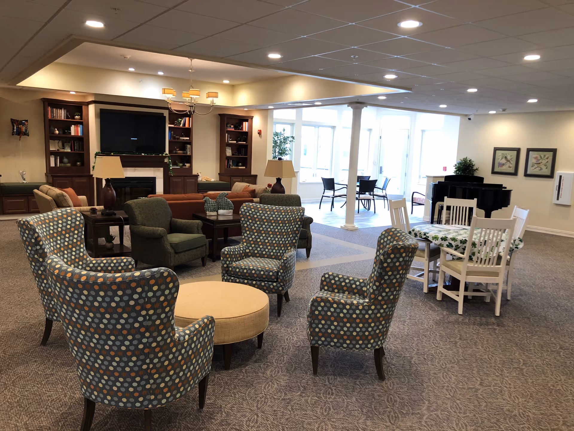 Spacious senior living common area with patterned armchairs arranged around a central ottoman, sofas by a fireplace and TV, a dining table, and a piano near large windows.