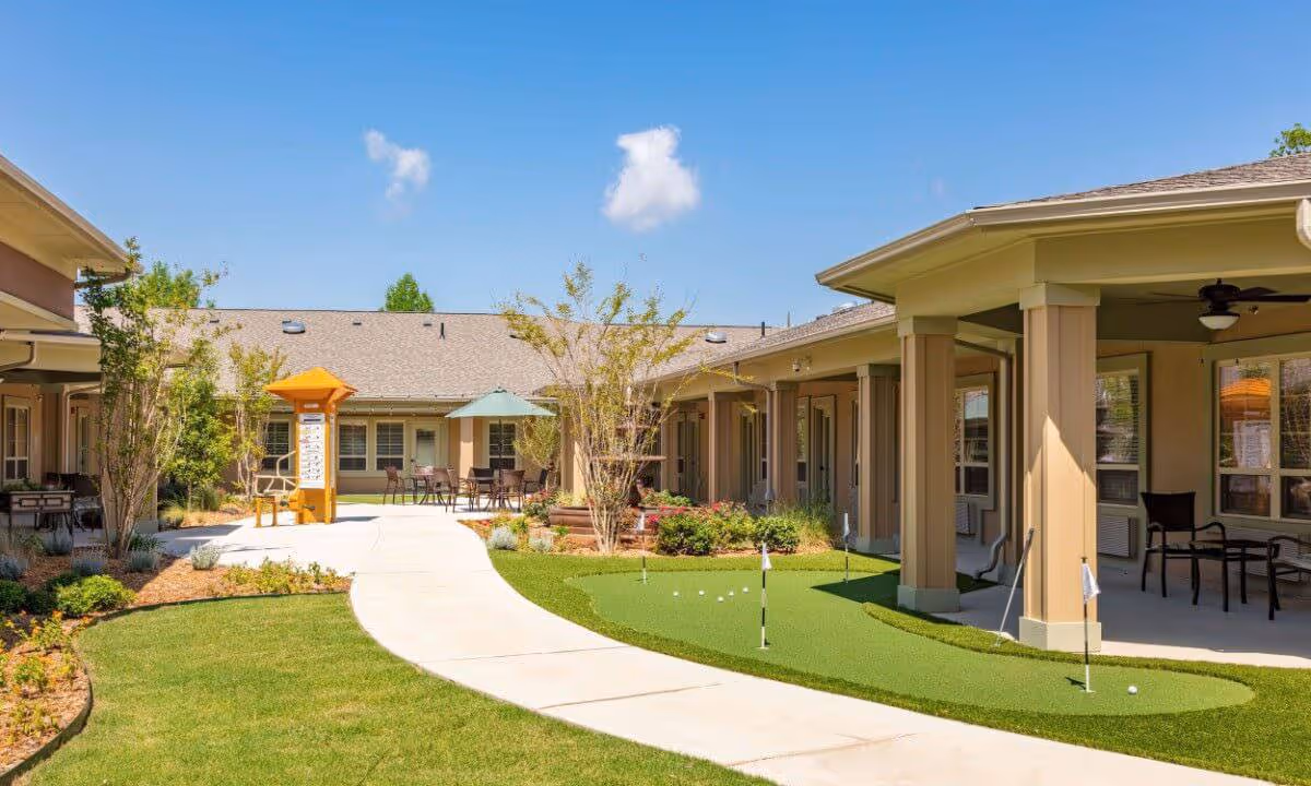 Sunny courtyard of a senior living community featuring a winding walkway, a small putting green, and covered patio seating.