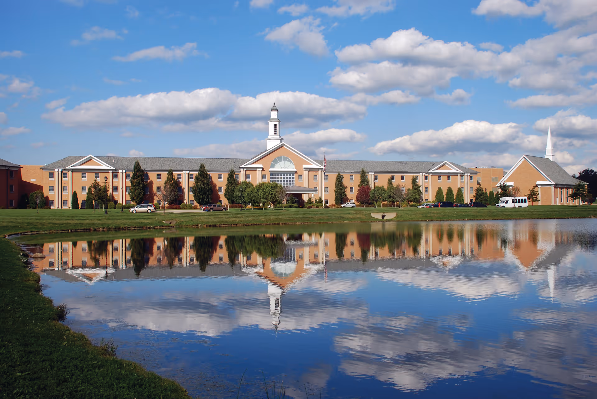 A large brick building with a central cupola and a smaller building with a steeple on the right, reflected in a calm pond in front. The sky is blue with scattered clouds, and there are trees and parked cars along the building.