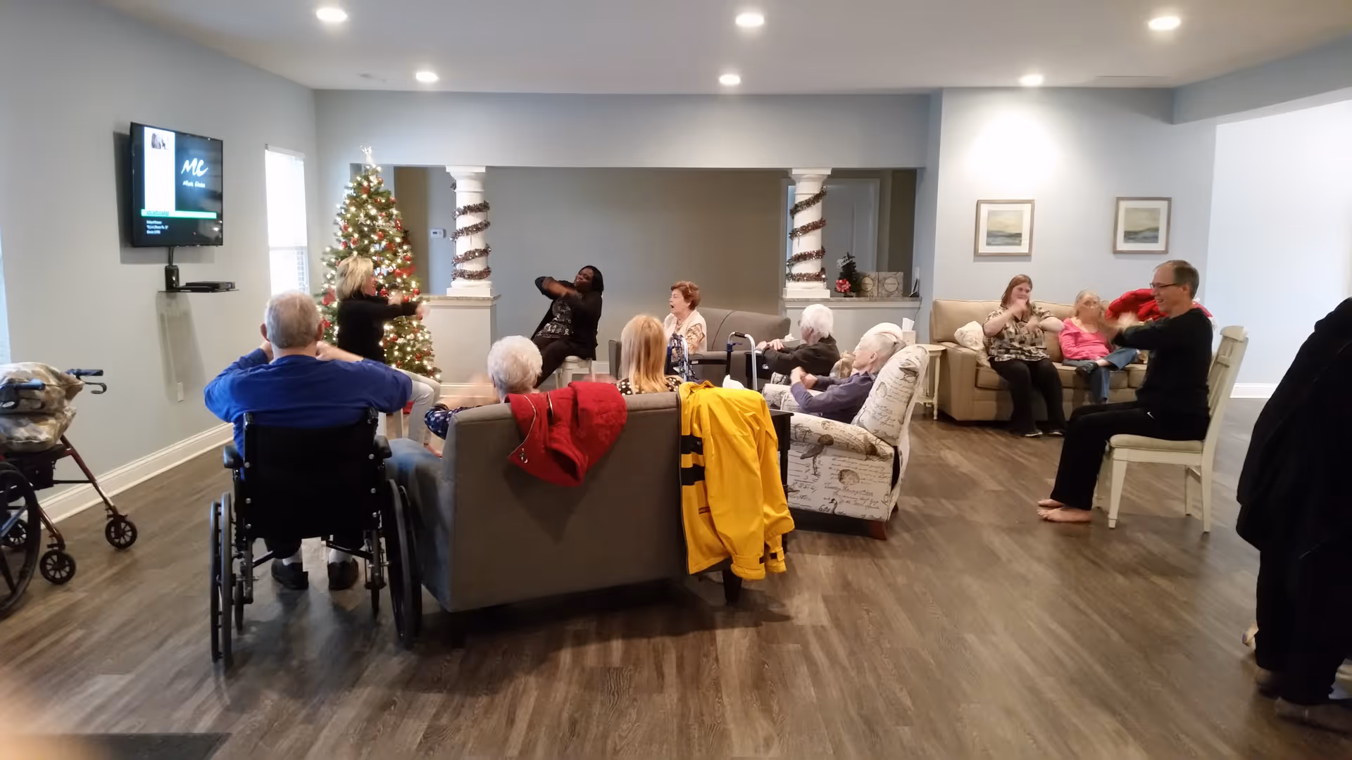 A group of elderly people and caregivers participating in a seated exercise session in a spacious living room decorated with a Christmas tree. The room has hardwood floors, comfortable seating including sofas and armchairs, and a wall-mounted TV.