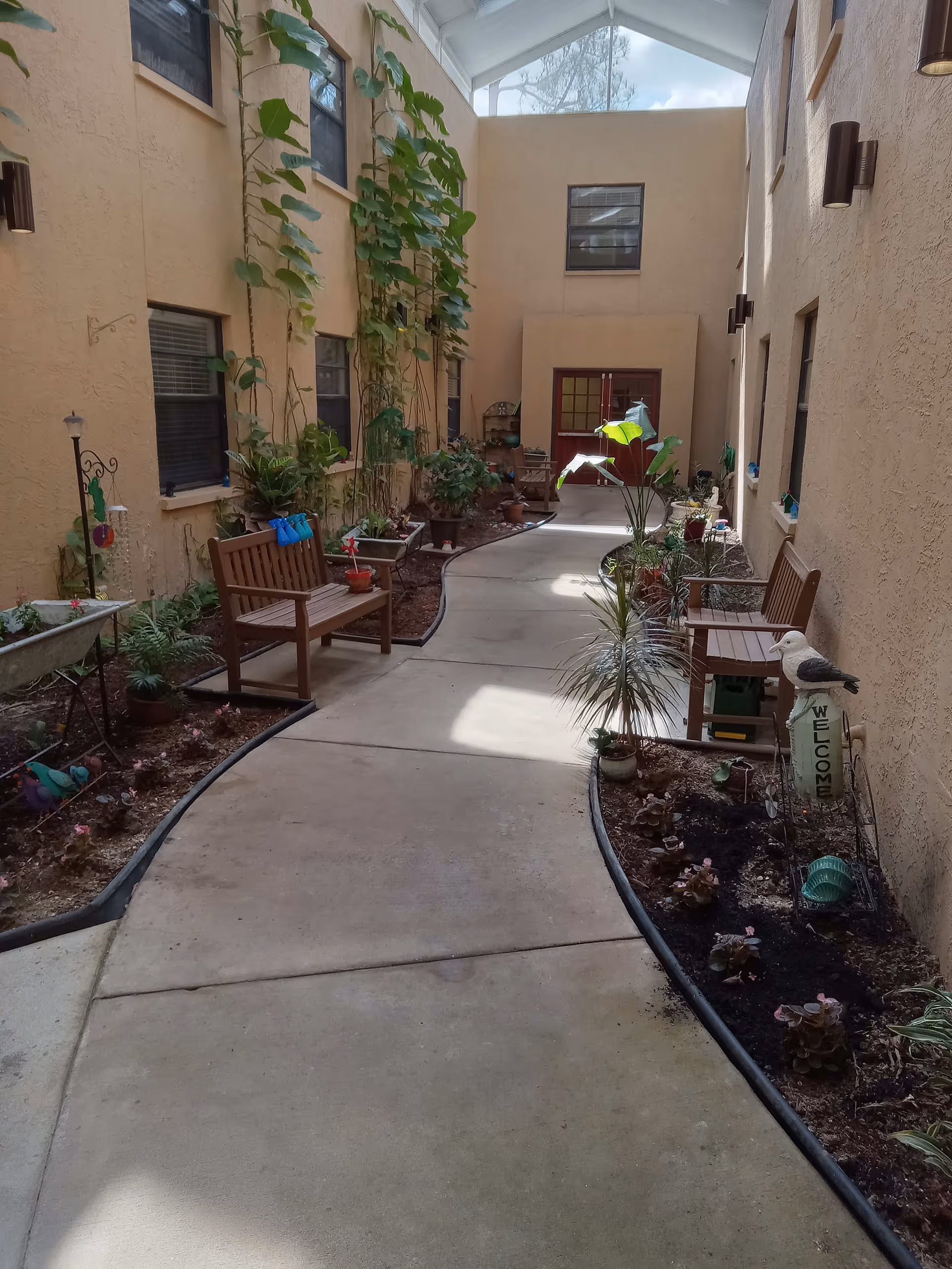 Indoor courtyard with a curved concrete walkway flanked by garden beds containing various plants and flowers. There are wooden benches along the walls, tall windows, and a glass ceiling allowing natural light to enter. A decorative bird statue holding a 'WELCOME' sign is visible on the right side.