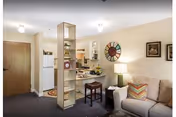 Interior view of a cozy living space in a retirement and assisted living facility featuring a beige couch with colorful pillows, a small wooden stool, a lamp on a side table, a decorative wall clock, and a kitchen area with white appliances and cabinets. A wooden door is visible in the background.