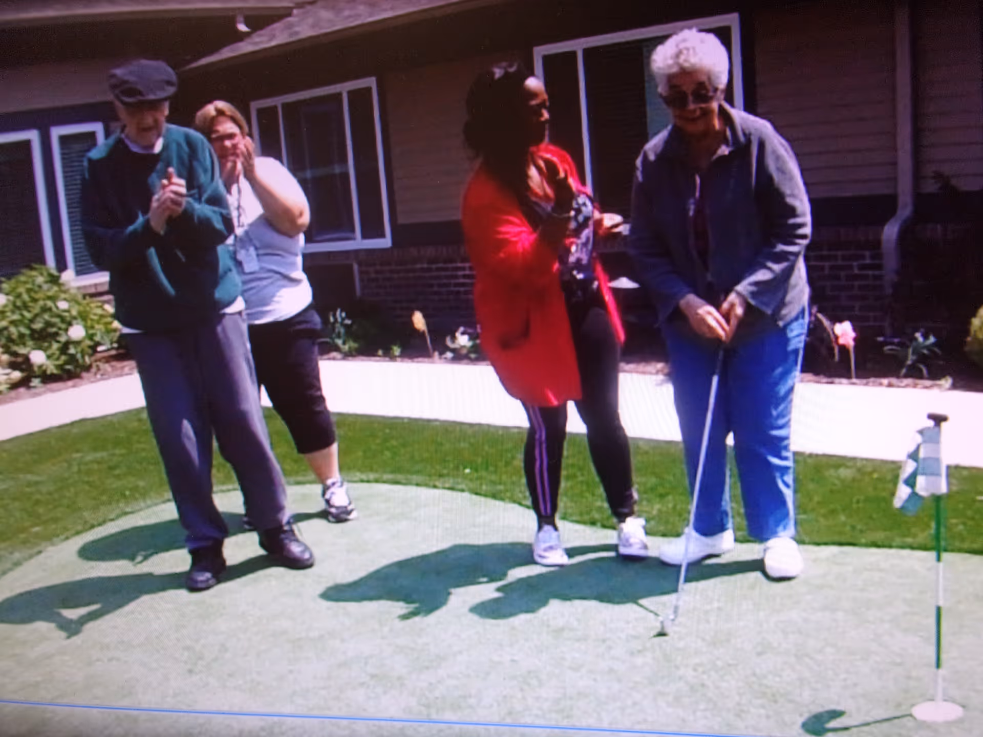 Four people standing on a putting green outside a building. One elderly woman is holding a golf putter preparing to putt, while the others watch and smile. The background shows a building with windows and some landscaping.