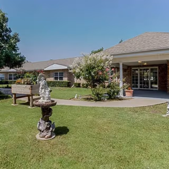 Front entrance of a single-story senior living building with lawn, garden statues, and a covered walkway.