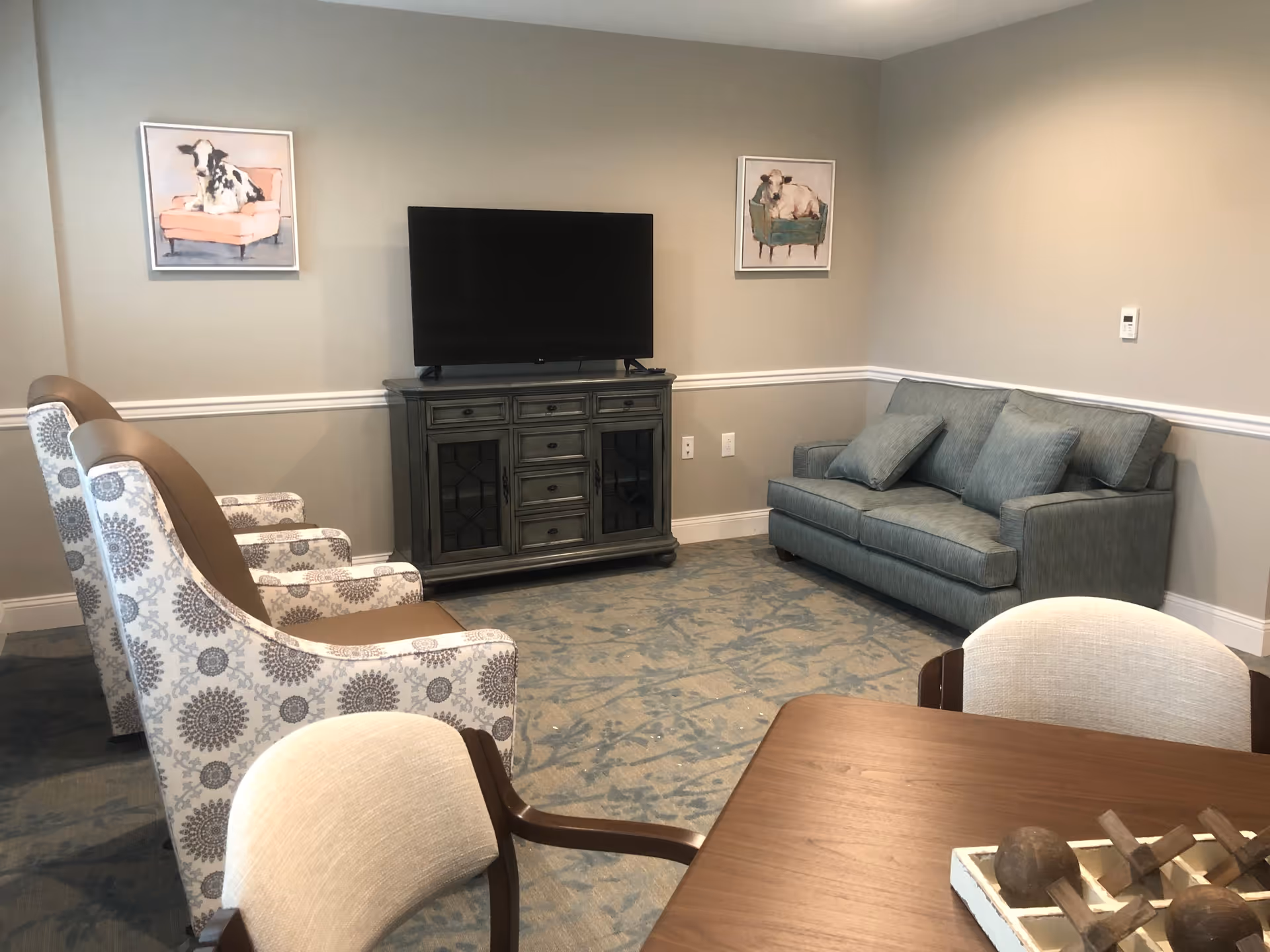 A cozy living room with a blue sofa, two patterned armchairs, a wooden TV stand with a flat-screen TV, and a wooden table with chairs. The walls are beige with white trim, and there are two paintings of cows sitting on chairs hanging on the wall.