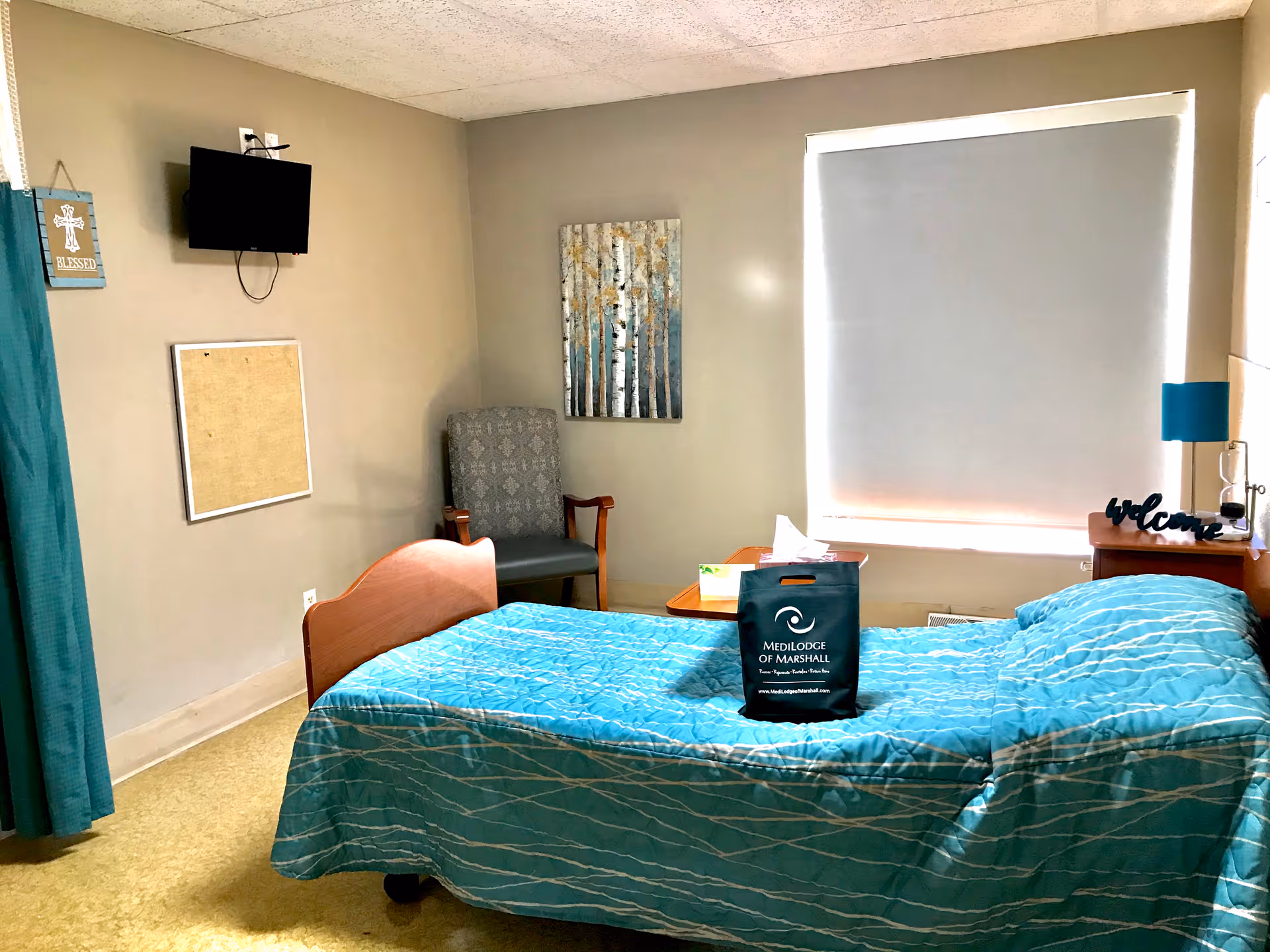 Small senior living bedroom with a single bed covered in teal bedding, a chair, wall-mounted TV, bedside table, and a Medilodge of Marshall bag on the bed.