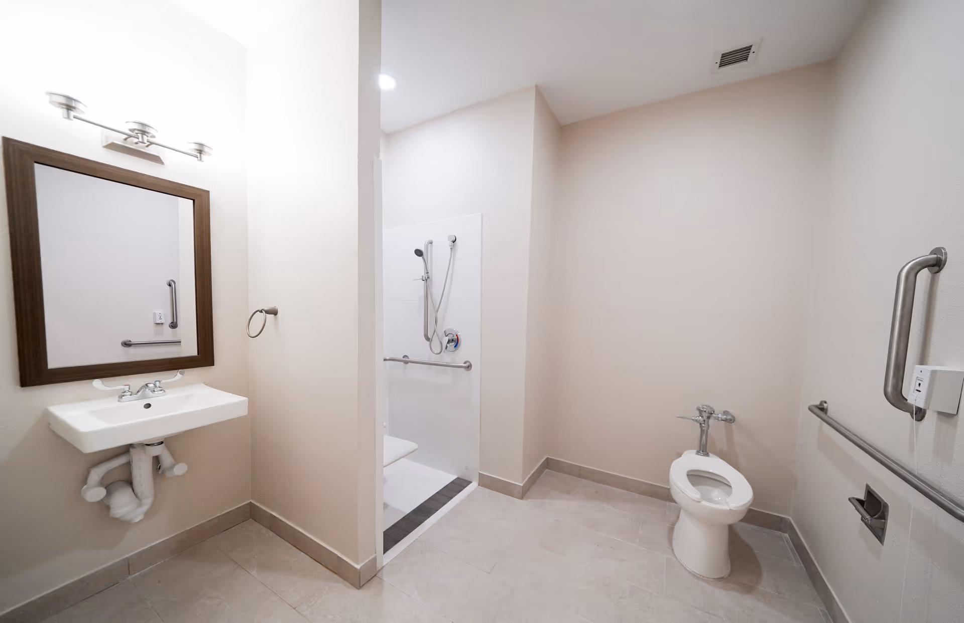 Accessible bathroom with a wall-mounted sink and mirror, a toilet with grab bars, and a roll-in shower with a handheld showerhead.