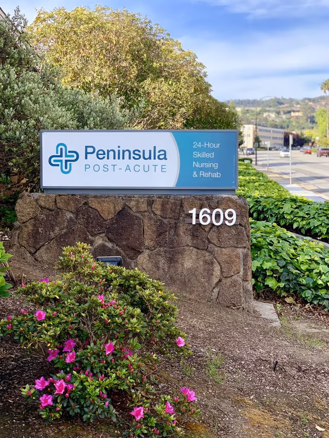 Outdoor stone sign for Peninsula Post-Acute facility with the address number 1609. The sign includes the text '24-Hour Skilled Nursing & Rehab' and is surrounded by greenery and pink flowers, with a road and buildings visible in the background under a blue sky.