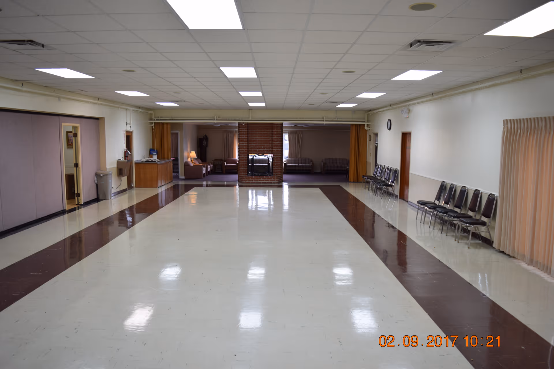 Empty multipurpose community room with a glossy tiled floor, chairs lined along the walls and a seating area visible at the far end.