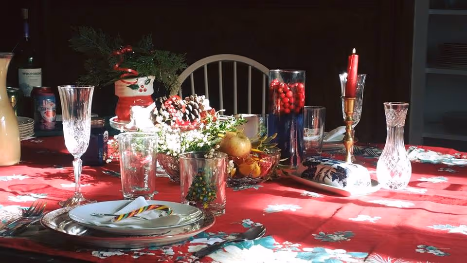 A dining table set with a red floral tablecloth, decorated with holiday-themed items including a candy cane on a plate, pine cones, berries, a red candle in a brass holder, and various glassware including wine glasses and water glasses.