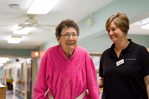 An elderly woman wearing a bright pink sweater is walking down a hallway with the assistance of a smiling female caregiver dressed in a black shirt with a name tag. The setting appears to be inside a senior living or rehabilitation facility.