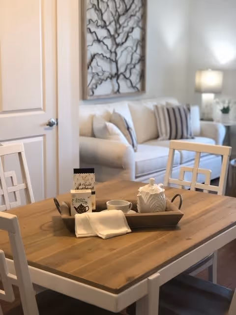 A wooden dining table with white chairs around it, featuring a tray with a white teapot, a cup, napkins, and a small packet. In the background, there is a beige sofa with striped pillows, a side table with a lamp, and a decorative wall art piece.