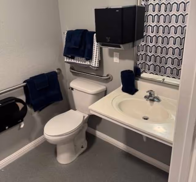A small bathroom with a white toilet and a white sink with a silver faucet. There are navy blue and white towels hanging on metal bars above and beside the toilet. A black paper towel dispenser is mounted on the wall above the toilet. A mirror with a patterned curtain reflection is visible above the sink. The walls are light gray and the floor is a darker gray.