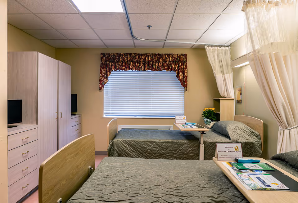 Shared care bedroom with two hospital-style beds, bedside tables, a wardrobe, and a curtained window.