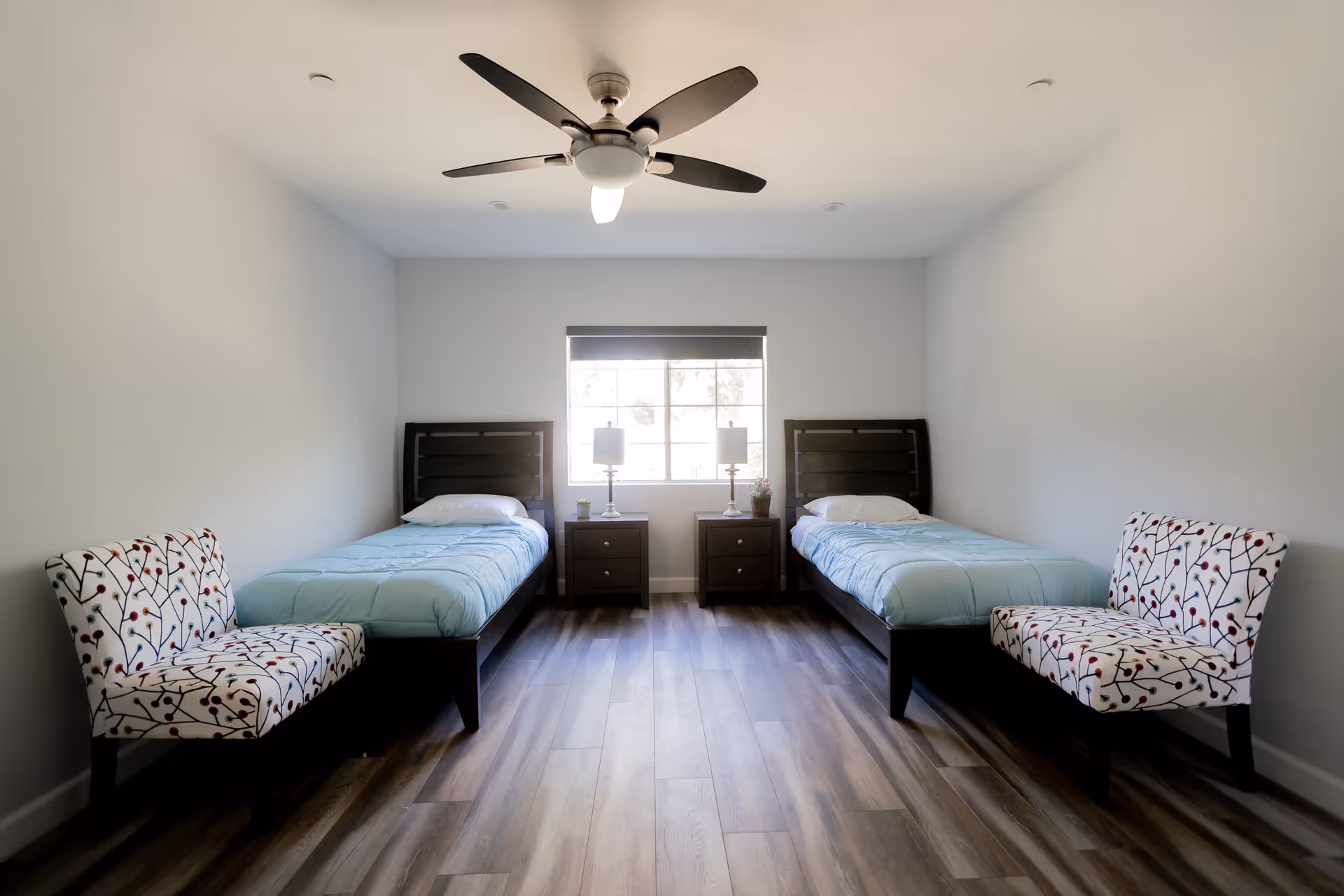 A simple bedroom with two single beds placed against opposite walls, each with a light blue comforter and white pillow. Between the beds is a window with a dark roller shade, flanked by two matching nightstands each holding a lamp and small plants. Two patterned chairs with a white background and colorful branch designs are placed at the foot of each bed. The room has wood flooring, white walls, and a ceiling fan with a light in the center of the ceiling.