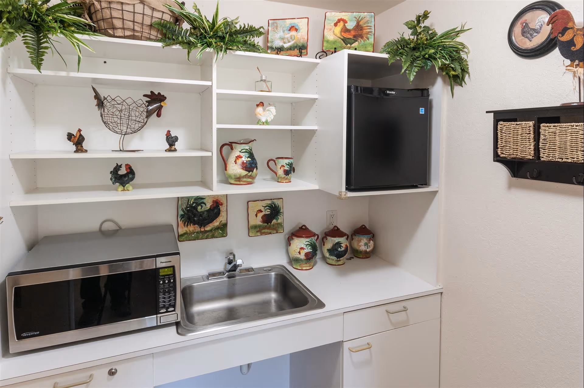 A small kitchenette area with a stainless steel sink, a microwave, and a mini refrigerator. White shelves above the sink hold various rooster-themed decorative items including figurines, pitchers, and plates. Green plants are placed on top of the shelves and a wall-mounted shelf with wicker baskets is visible on the right side.