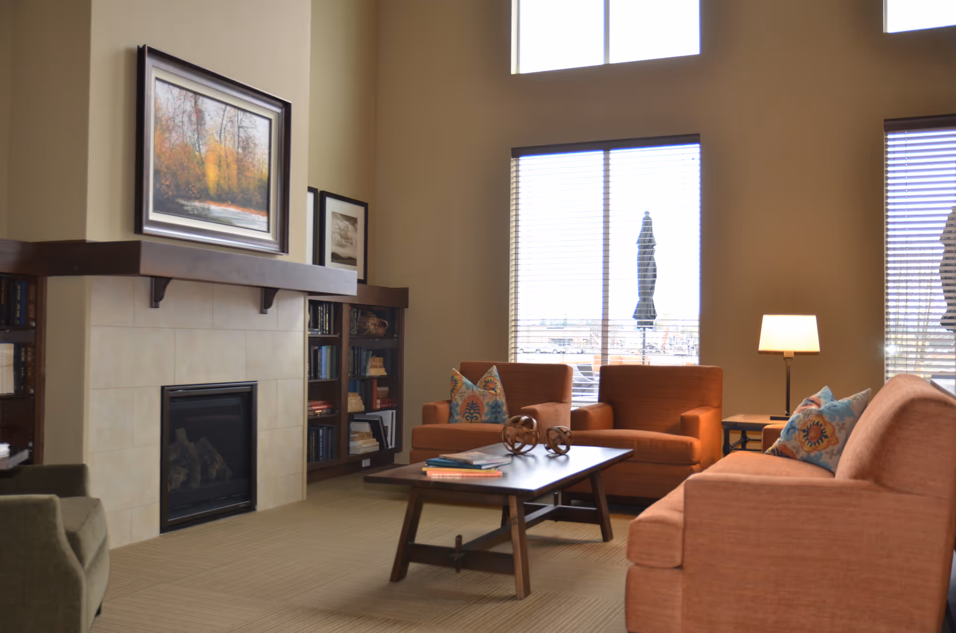 A cozy living room with beige walls and large windows letting in natural light. The room features a fireplace with a wooden mantle, bookshelves on either side, and framed artwork above the fireplace. There are two orange armchairs and an orange sofa with colorful patterned pillows arranged around a wooden coffee table with decorative items and books. A floor lamp is placed next to one of the armchairs.
