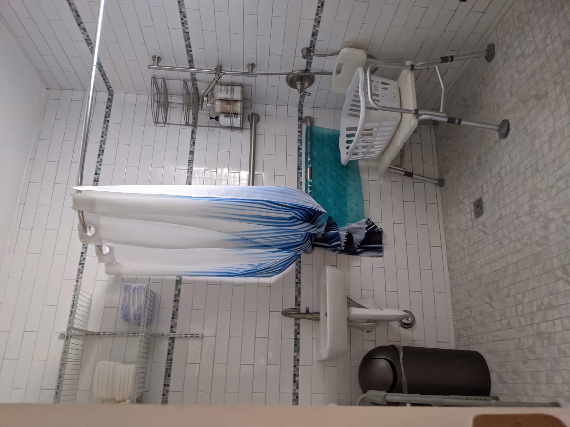 A bathroom with white tiled walls and a tiled floor. There is a small wall-mounted sink with a faucet, a trash can, and a shower area with a white and blue shower curtain. Inside the shower area, there is a white shower chair with a laundry basket on it, a grab bar, a showerhead, and a metal rack holding bottles of soap or shampoo. Towels are neatly stacked on a wire shelf mounted on the wall.