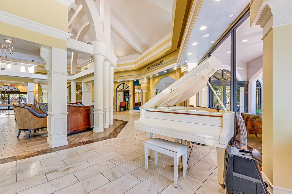 Bright and spacious senior living facility common area with a white grand piano and matching bench near large windows. The room features high vaulted ceilings with architectural columns, comfortable seating areas with sofas and chairs, and a sign indicating the North Tower. The floor is tiled with a mix of light and dark tiles, and natural light fills the space.