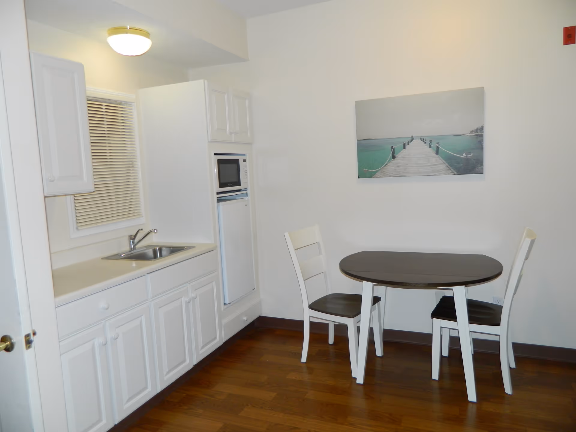 Small kitchen area with white cabinets, a sink, a microwave, and a mini refrigerator. Adjacent to the kitchen is a small dining area with a round table and two chairs. A wall-mounted picture of a wooden pier extending into turquoise water is visible above the table.
