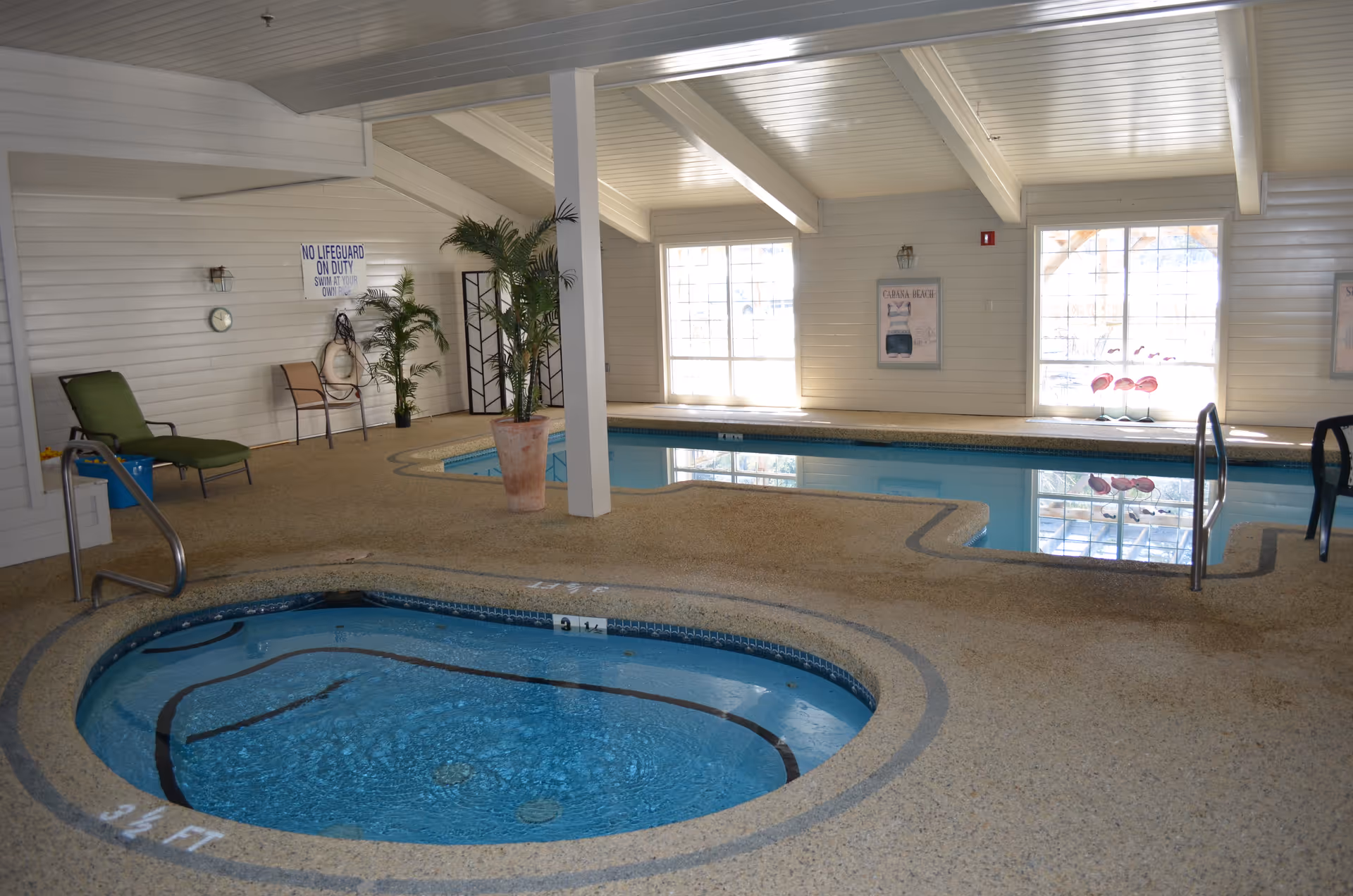 Indoor pool area with a small hot tub in the foreground and a larger swimming pool in the background. The room has white paneled walls and ceiling with large windows letting in natural light. There are a few chairs and potted plants around the pool area, and a sign on the wall reads 'No Lifeguard On Duty Swim At Own Risk.'