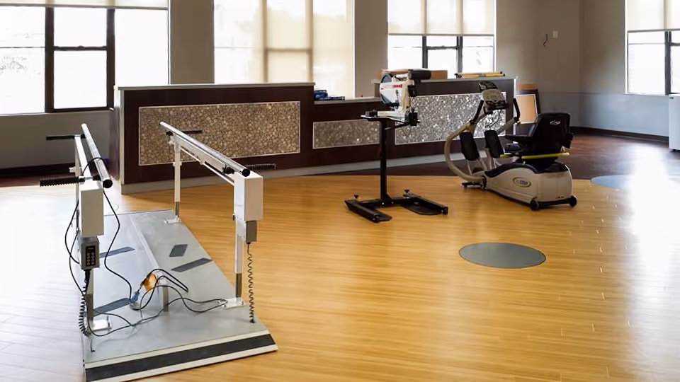 A rehabilitation room with exercise equipment including parallel bars for walking practice and a recumbent exercise bike, set on a wooden floor with large windows in the background.