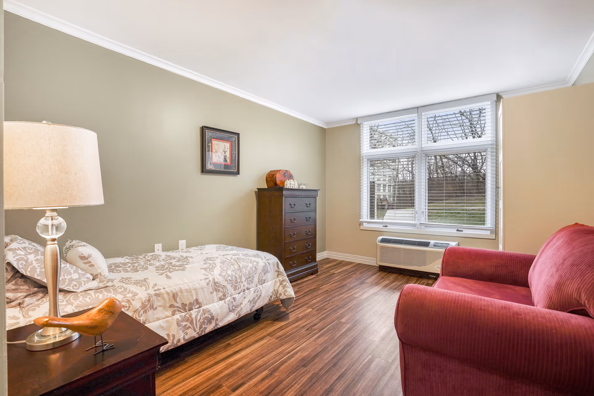 A cozy bedroom with a single bed covered in a patterned beige and white comforter, a wooden nightstand with a lamp and a decorative bird figurine, a dark wooden chest of drawers, a large window with white blinds, and a red upholstered armchair on wooden flooring.