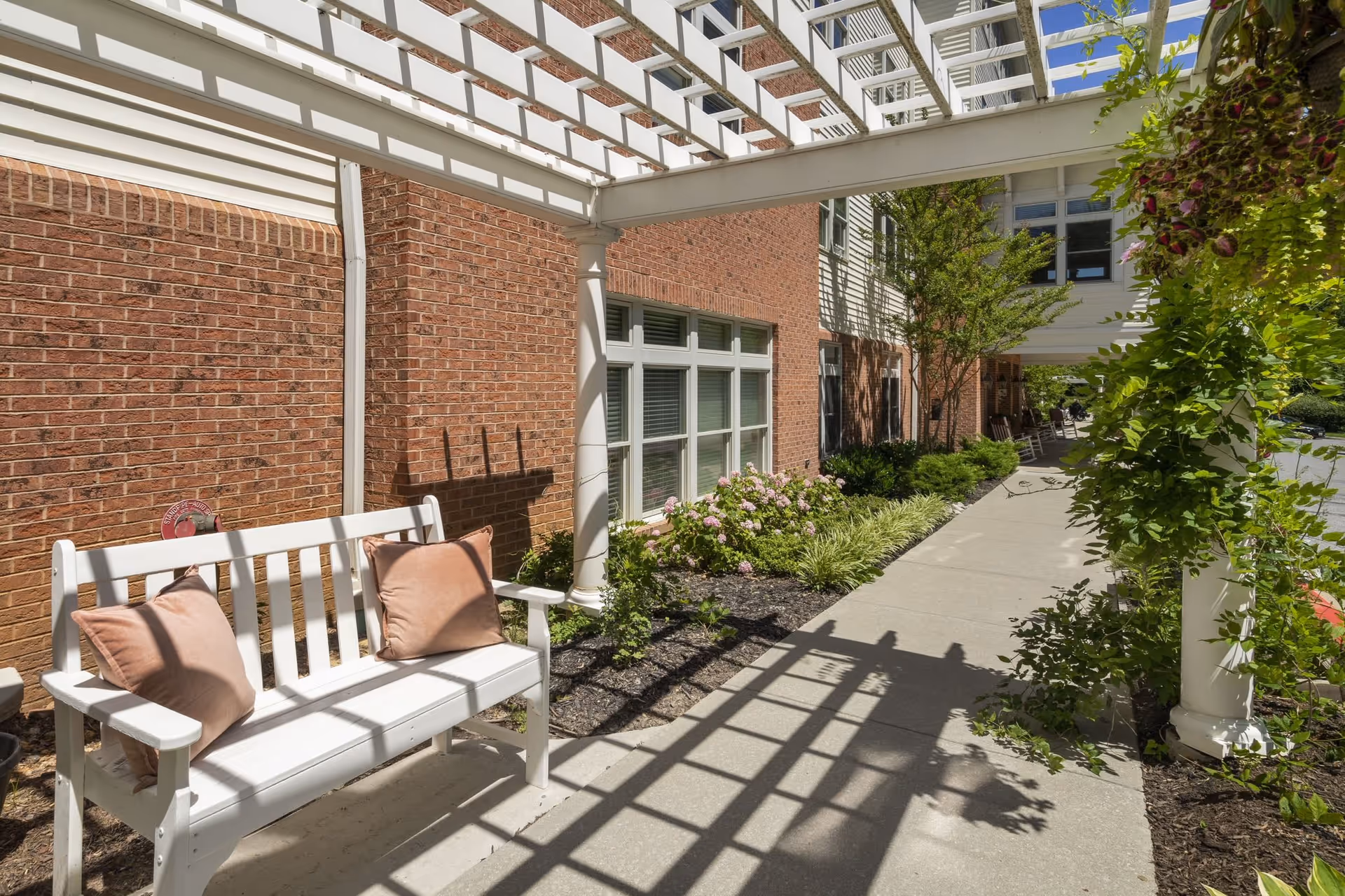 Outdoor walkway alongside a brick building with a white pergola casting shadows. A white bench with two brown cushions is placed near the building, surrounded by green plants and flowers.