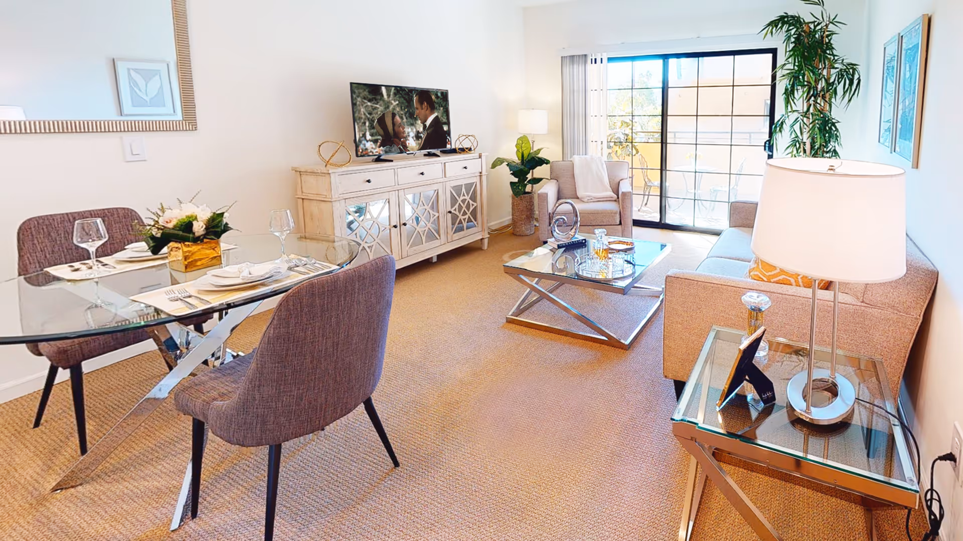 A bright and modern living room and dining area with beige carpet. The dining area has a glass table with four purple upholstered chairs and place settings with wine glasses. The living room features a beige sofa, an armchair, a glass coffee table with decorative items, a side table with a lamp, and a white cabinet with a TV on top. Large sliding glass doors lead to an outdoor balcony with plants visible outside.
