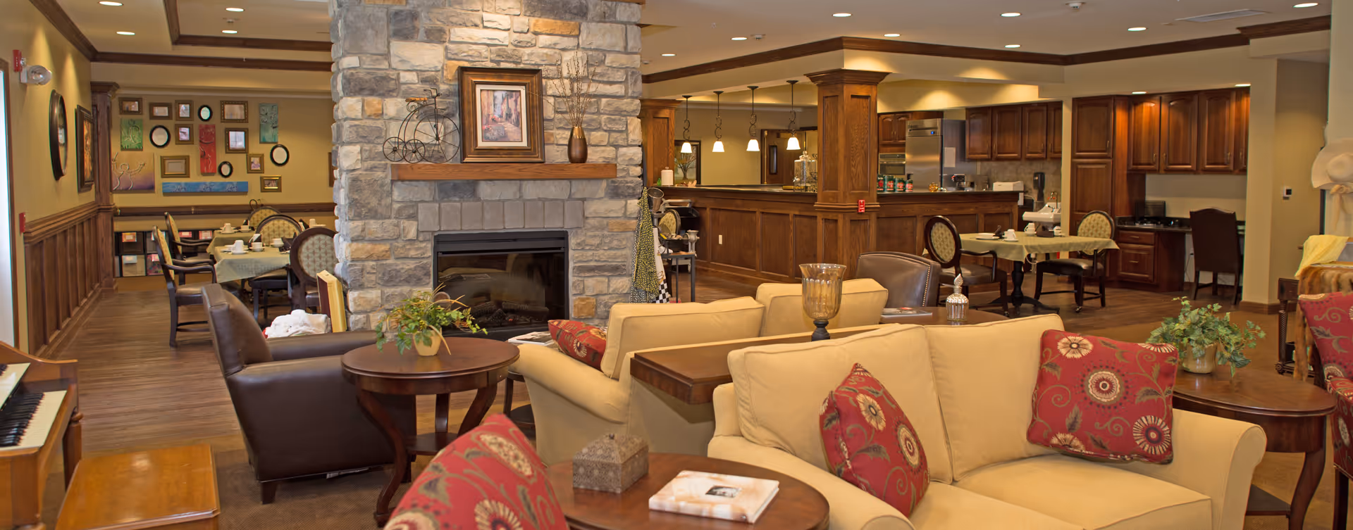 A cozy senior living facility common area featuring a stone fireplace with a framed picture and decorative items on the mantel. Surrounding the fireplace are comfortable beige sofas with red patterned pillows, wooden side tables with plants and decorative objects, and leather chairs. In the background, there is a dining area with tables and chairs, a kitchen with wooden cabinets and stainless steel appliances, and warm lighting throughout the space.