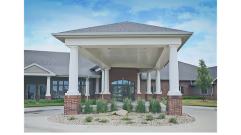 Front exterior view of a single-story brick building with a covered entrance supported by white columns. There is landscaping with rocks and plants in front of the entrance, and the sky is partly cloudy.