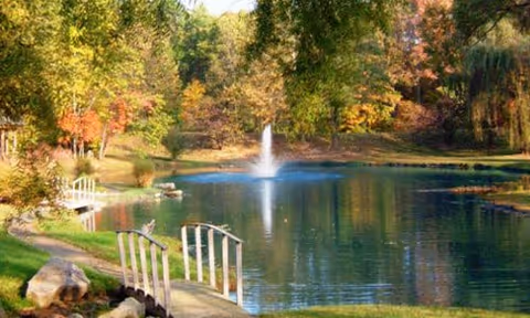 Small lake with a central fountain, grassy banks, arched wooden footbridges, and trees showing autumn foliage.