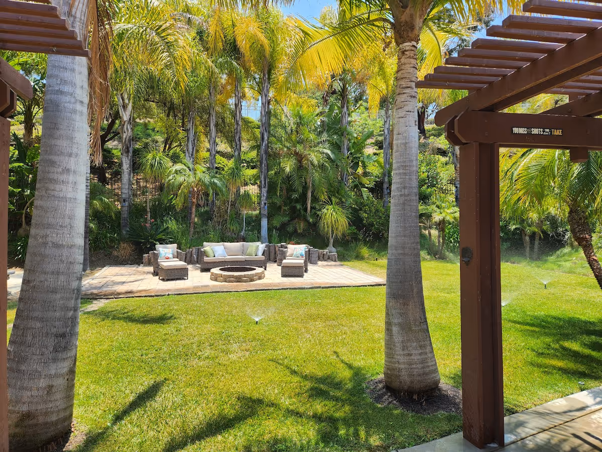 Outdoor garden area with green grass, palm trees, and a seating area consisting of a curved sofa and two chairs around a circular fire pit. The area is shaded by a wooden pergola and surrounded by lush tropical plants and trees.