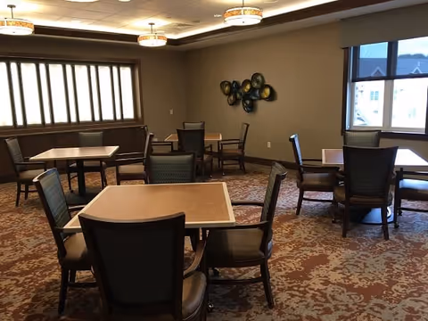 A dining room with several square tables and cushioned chairs arranged around them. The room has patterned carpet flooring, beige walls, and two windows letting in natural light. Decorative wall art is visible on one wall, and ceiling lights provide additional illumination.