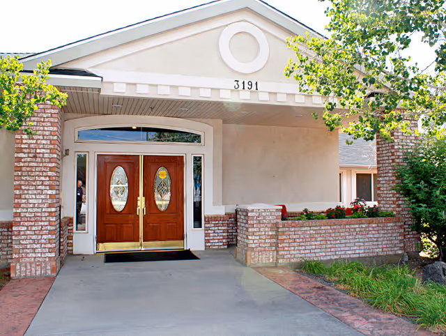 Entrance to a building with double wooden doors featuring oval glass panels, brick pillars on either side, and a covered porch area with the number 3191 above the doors. There are green trees and plants around the entrance.
