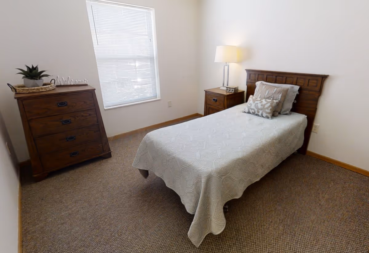 A simple, neatly arranged bedroom with a single bed covered in a light-colored quilt and decorative pillows. There is a wooden headboard, a wooden nightstand with a lamp and a small decorative item, and a wooden dresser with a plant and a 'Welcome' sign. A window with blinds allows natural light into the room.