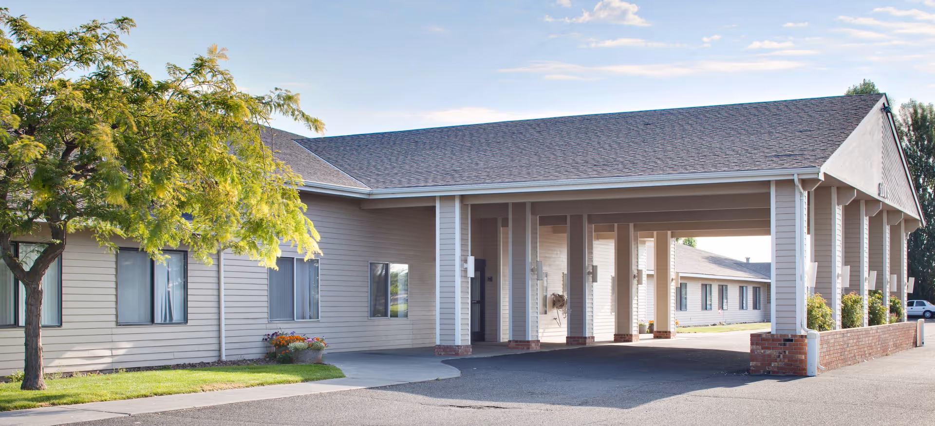 Exterior view of Columbia Crest Center showing a single-story building with beige siding, a covered entrance supported by white columns, a tree with green leaves on the left, and a clear sky with some clouds.