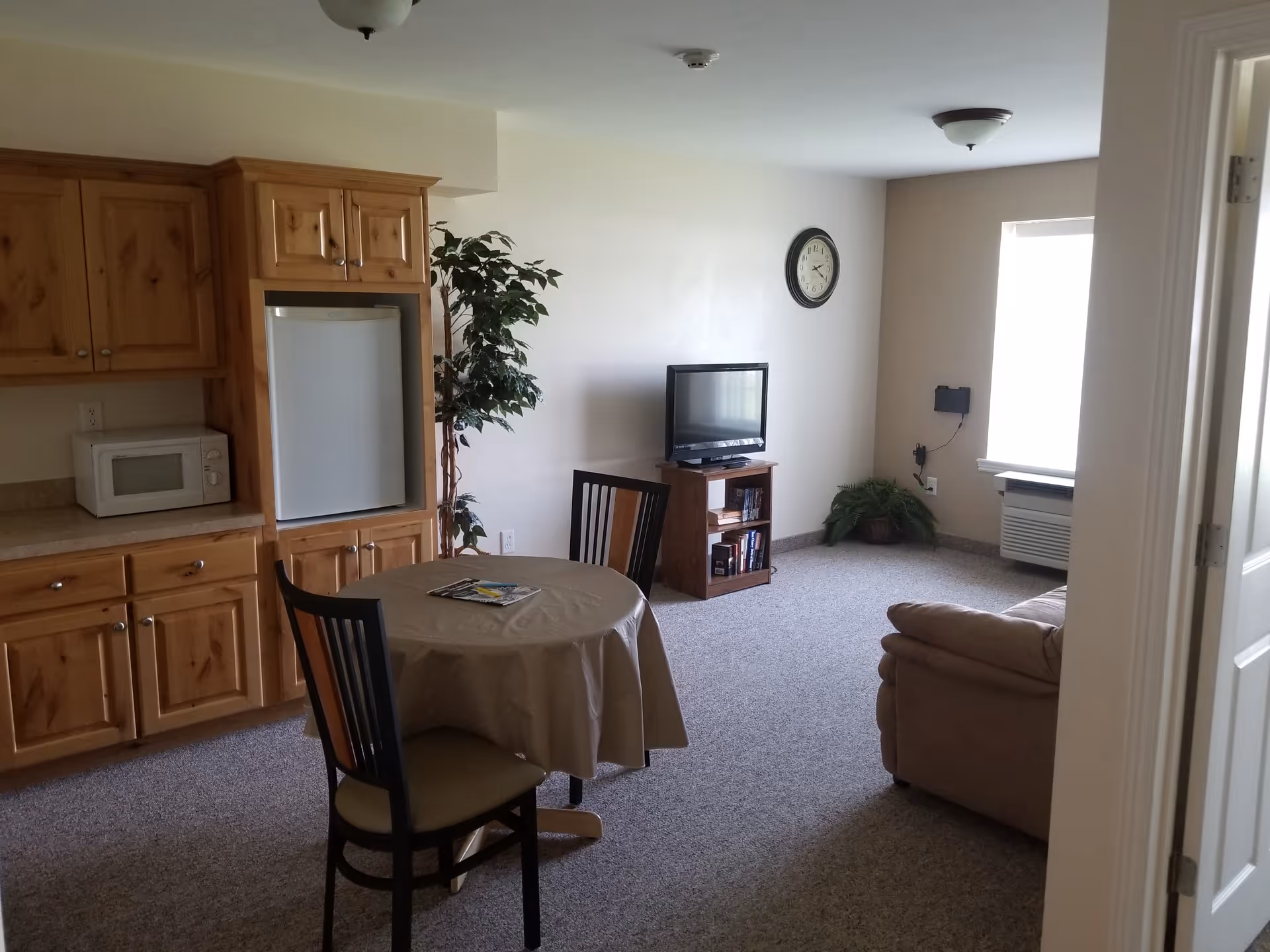 Assisted living apartment living area with a round table and chairs, kitchenette cabinets and microwave, a TV on a stand, and a sofa by a window.