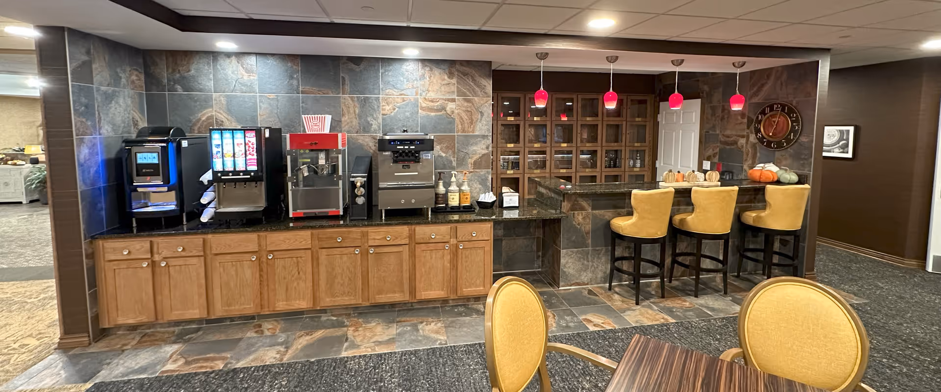 A beverage and snack station area in a retirement community featuring a coffee machine, soda dispenser, popcorn machine, and condiment bottles on a granite countertop with wooden cabinets below. To the right, there is a bar counter with three yellow cushioned bar stools, red pendant lights hanging above, a wall clock, and decorative pumpkins. In the foreground, there is a table with yellow cushioned chairs.