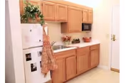 Small kitchenette with light wood cabinets, white countertops, sink, microwave and a refrigerator with an apron hanging on it.