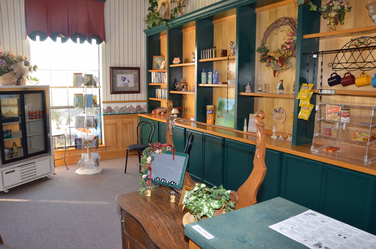 Interior view of a small shop or common area with wooden shelves displaying various decorative items, snacks, and drinks. There is a small refrigerator on the left side, a rack with greeting cards near a window with a red valance, and a wooden counter with chairs. The room has a cozy and welcoming atmosphere with plants and a small chalkboard sign that says 'Welcome'.