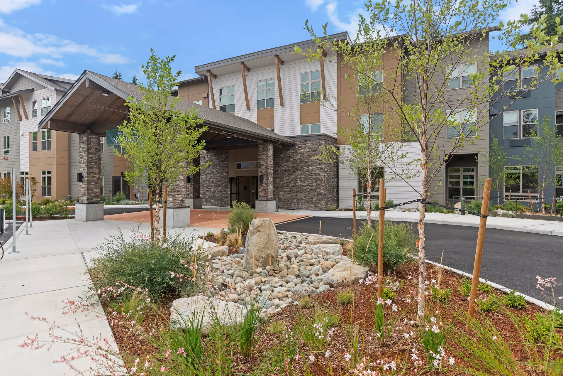 Entrance of a modern senior living facility with a covered drop-off area, surrounded by landscaped greenery including small trees, shrubs, and flowering plants. The building has a mix of stone and siding exterior with multiple windows and a clear blue sky above.