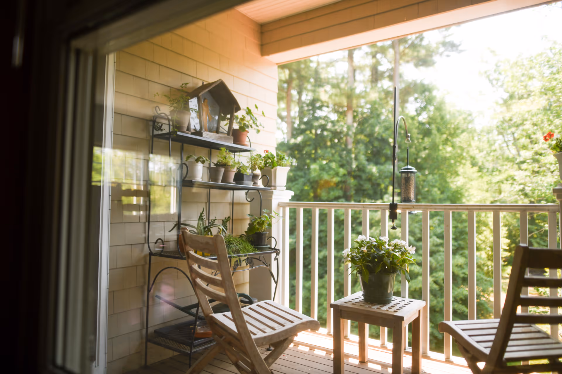 A cozy outdoor balcony with wooden chairs and a small table holding a potted plant. There is a metal shelf with multiple potted plants against the wall, and a bird feeder hanging from the ceiling. The balcony overlooks a lush green forested area.