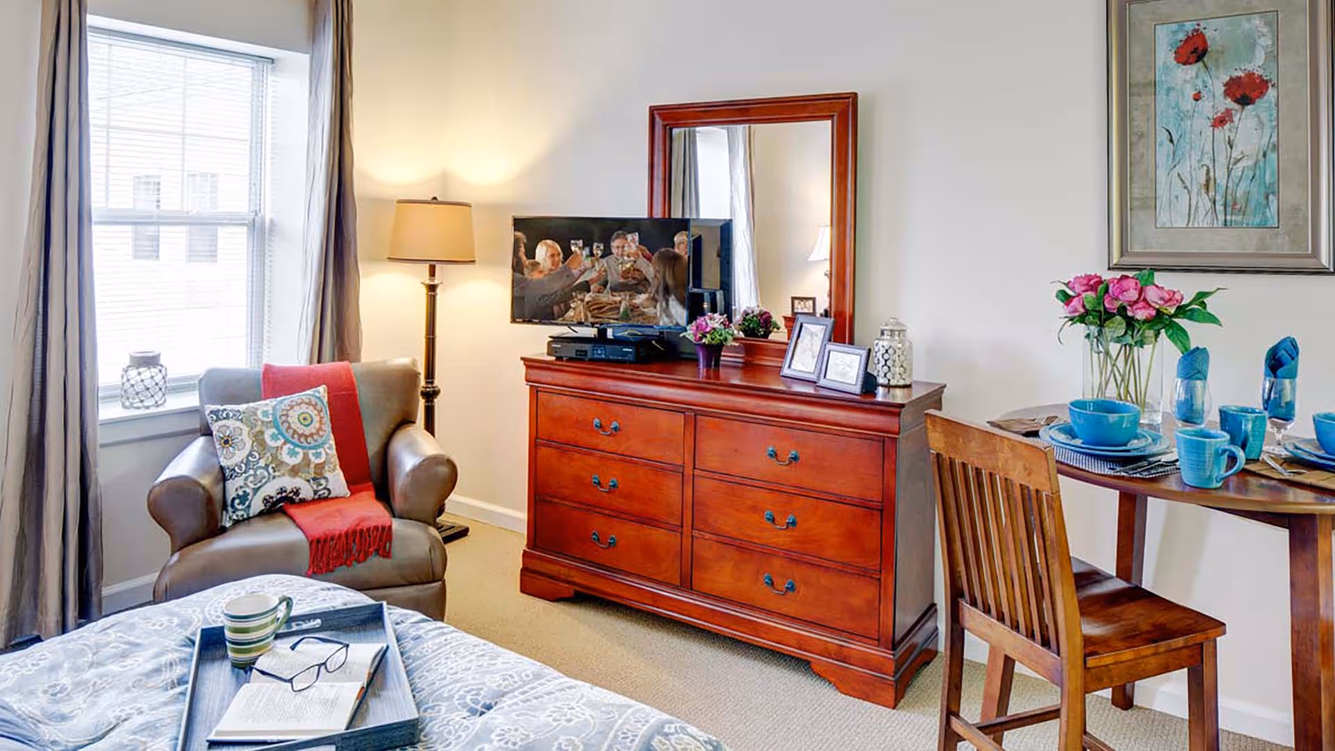 A cozy senior living room with a leather armchair draped with a red throw and a patterned pillow next to a window with curtains. A wooden dresser with a large mirror holds a flat-screen TV, framed photos, and decorative items. A small wooden dining table set with blue dishes and napkins is positioned against the wall, which is adorned with a floral painting. A floor lamp provides warm lighting.